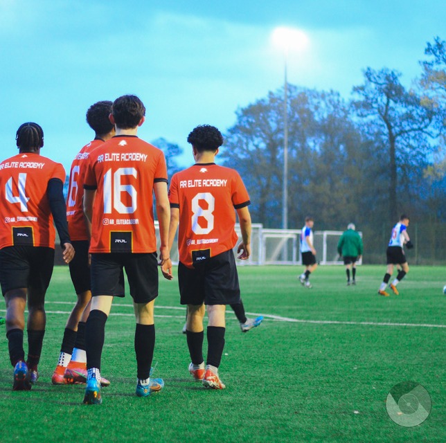 AR Elite Academy players in orange shirts walking off the pitch under floodlights at dusk